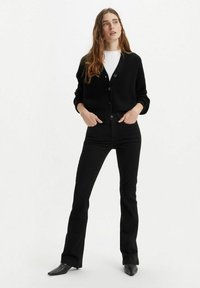 Zwarte gebreide cardigan met ribbeltextuur, voorzien van knopen aan de voorkant. Gepaarde met zwarte high-waisted flared jeans en schoenen met puntige neuzen.