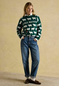 Grüner Strickpullover mit weißem Schafsmuster und dem Text "Fleece Navidad", kombiniert mit blauen Jeans, die an den Cuffs aufgerollt sind, und braunen Loafers.