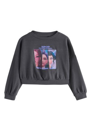 Junges Mädchen, das locker sitzende beige Hosen, weiße Sneakers und ein graues Sweatshirt mit K-Pop Demon Hunters Charaktermotiven trägt.