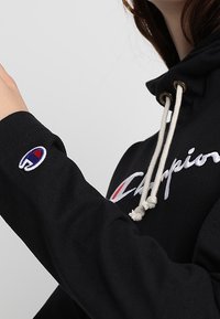 Zwarte hoodie met een trekkoord, met witte geborduurde "Champion" tekst en een kleurrijke logopatch op de linker mouw.