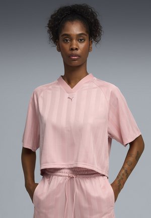 Femme aux cheveux bouclés portant un haut court à rayures roses claires de la marque Puma et un short assorti, debout les mains dans les poches.