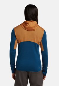 Persona con cabello largo y oscuro que lleva puesta una chaqueta con capucha en tonos azul y naranja, de espaldas contra un fondo blanco.