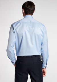 Camicia azzurro chiaro a maniche lunghe con una texture liscia e un colletto classico. Pantaloni scuri con tasche decorative e una cintura elegante.