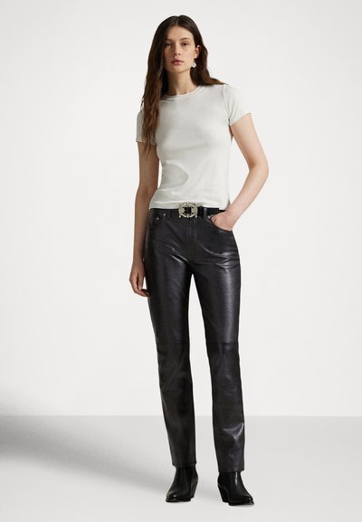 Γυναίκα που φοράει λευκό πουκάμισο με κοντά μανίκια, μαύρο δερμάτινο παντελόνι με διακοσμητική ζώνη και μαύρα μποτάκια αστραγάλου, στέκεται μπροστά σε μια απλή φόντο.