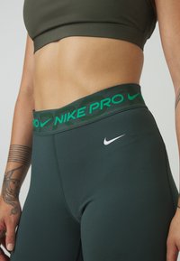 Nike Pro legíny ve tmavě zelené barvě s hladkou texturou. Mají široký zelený pás s nápisem "NIKE PRO" a bílým logem Nike.