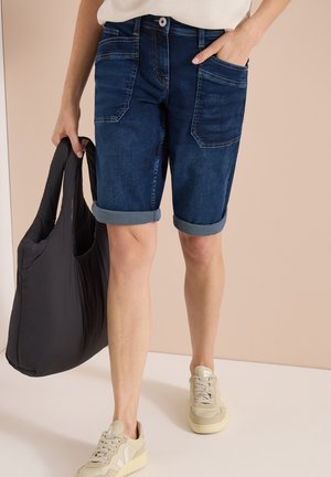 Person iført opsmøgede mørke denimshorts, beige sneakers, holder en stor sort taske i den ene hånd, den anden hånd i lommen.