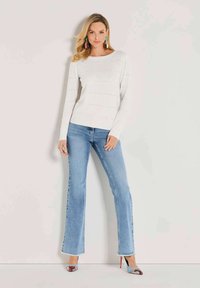 Frau in einem weißen Langarmpullover, hellblauen Schlagjeans, bunten High Heels und großen, mehrfarbigen Ohrringen, die vor einer schlichten Wand steht.
