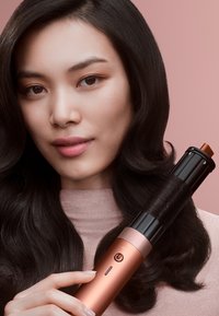 Piastra arricciacapelli con un tubo nero lucido e un manico oro rosa che presenta un indicatore di accensione; l'utente la tiene accanto ai suoi capelli fluenti.