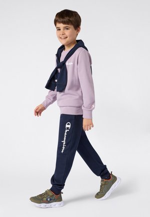 Sudadera color lila claro con texto "Champion", pantalones azul marino con estampado de logo y zapatillas verdes con suelas blancas y diseño detallado.