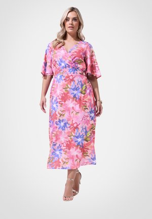 Vrouw in een roze midi-jurk met blauwe en paarse bloemen, korte mouwen, V-hals en hakken met enkelriem die vooruit loopt.