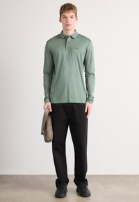 Long-sleeve green polo shirt features a collar and subtle logo, paired with black trousers and dark sneakers, holding a beige shoulder bag.