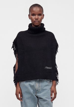 Personne portant un poncho noir à franges avec un col roulé par-dessus un jean bleu clair, debout devant un fond blanc uni.