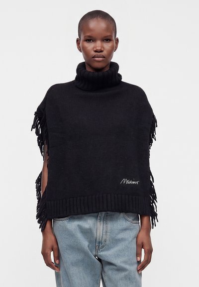 Personne portant un poncho noir à franges avec un col roulé par-dessus un jean bleu clair, debout devant un fond blanc uni.