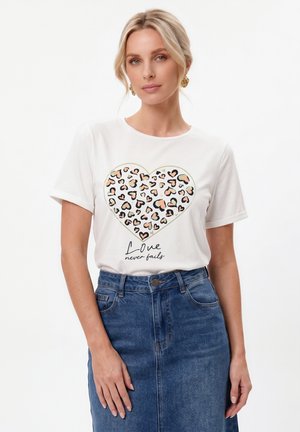 Blondynka w białej koszulce z nadrukiem serca w panterkę i tekstem "Love never fails,", w zestawieniu z niebieską spódnicą jeansową.