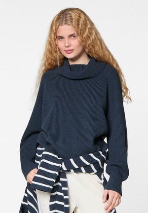 Jonge vrouw met lang, krullend blond haar, gekleed in een donkerblauwe geribde trui en een gestreepte marineblauwe en witte trui om haar middel geknoopt.