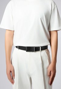 Svart läderbälte med en strukturerad yta och ett spänne i silvermetall, bärs över vita byxor och en vit t-shirt.