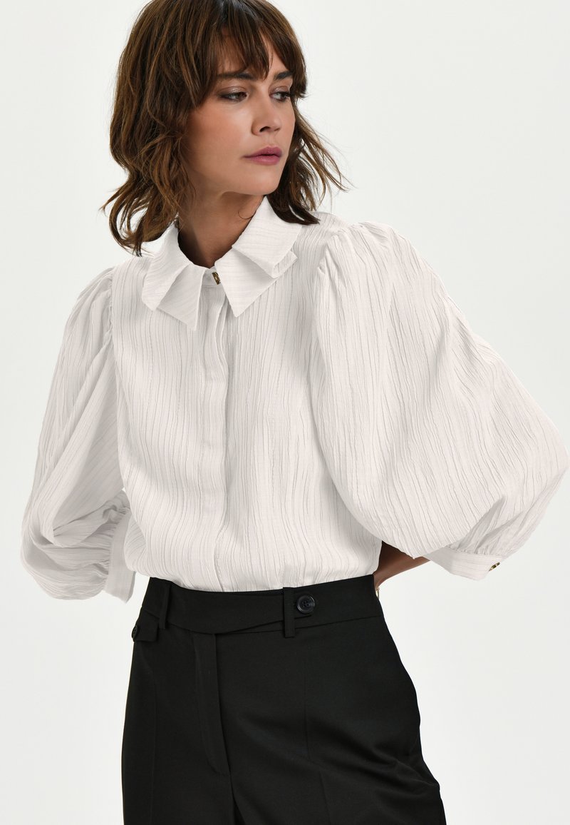 Weißes, strukturiertes Blusenshirt mit Kragen und Puffärmeln, kombiniert mit hochgeschnittenen schwarzen Hosen mit seitlichen Knopfdetails.