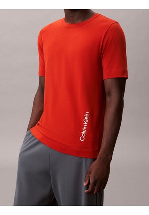Mężczyzna w jednolitej pomarańczowej koszulce z krótkim rękawem z logo „Calvin Klein” w pobliżu dołu, w połączeniu z szarymi spodniami.