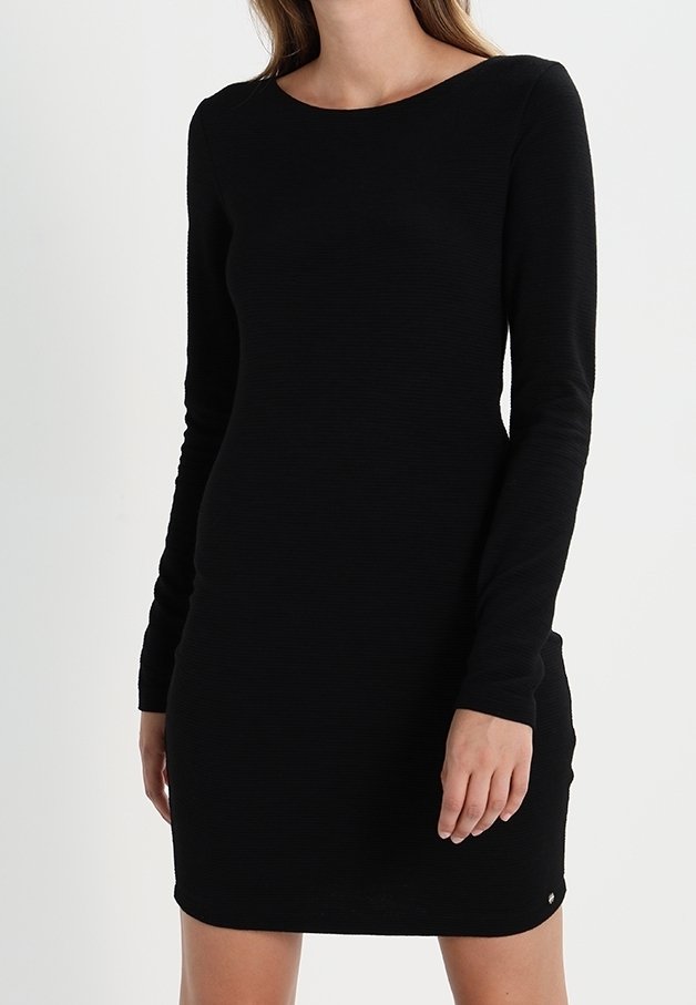 Femme portant une robe noire moulante à manches longues avec un col rond, debout devant un fond blanc uni.
