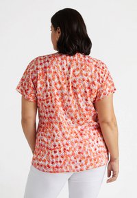 T-shirt à manches courtes en tissu léger, arborant un motif abstrait orange et rouge. Le design comprend un ourlet arrondi et une coupe décontractée.