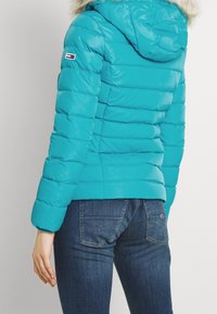 Turquoise donsjack met capuchon, voorzien van horizontale doorgestikte lijnen en een bontkraag. Mouw met een klein logo-embleem. Gecombineerd met een blauwe spijkerbroek.