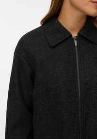 Femme portant une veste en laine gris foncé à fermeture éclair avec un col replié, visible de l'épaule au menton sur un fond blanc uni.