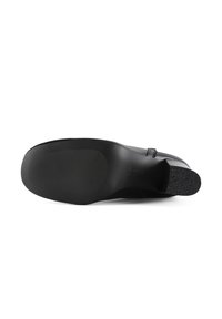 Zwarte leren schoen met een gestructureerde rubberen zool, gebogen vorm en zijgesp detail; de zool heeft een ribbelpatroon voor grip.