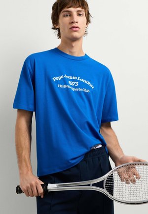 Junger Mann trägt ein blaues Pepe Jeans London T-Shirt und hält einen silbernen Tennisschläger vor weißem Hintergrund.