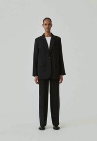 Black blazer with a single button closure, two front pockets, paired with straight black trousers and a white t-shirt. Smooth texture.