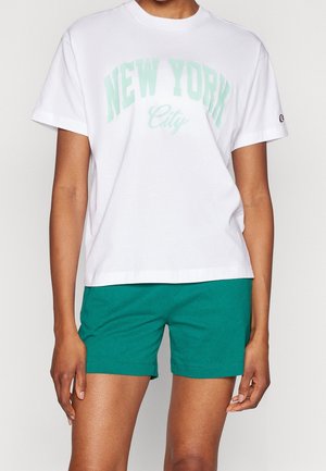 Biała koszulka z krótkim rękawem z napisem "NEW YORK City" w jasnym zielonym kolorze, zestawiona z zielonymi casualowymi szortami na stojącej osobie.