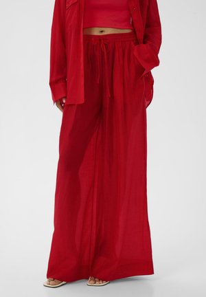 Kobieta w luźnych, czerwonych spodniach o szerokich nogawkach z troczkiem w pasie, czerwonym krótkim topie i pasującej czerwonej koszuli, stojąca z jedną ręką w kieszeni.