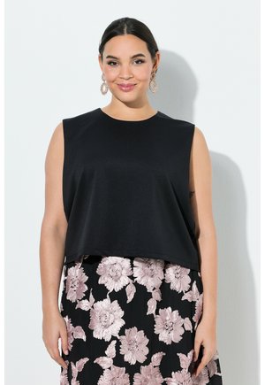 Kvinde med mørkt hår iført en ærmeløs sort top og sort nederdel med store lyserøde blomsterprint, smilende mod en hvid baggrund.