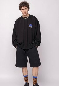Black oversized sweatshirt with blue graphic and text, paired with black shorts. The model wears brown leopard print socks with blue accents.
