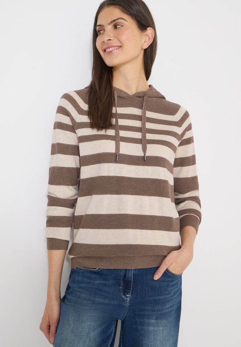 Femme portant un sweat à capuche rayé marron et beige avec cordons, les mains dans les poches, regardant sur le côté, vêtue d'un jean bleu sur un fond blanc.