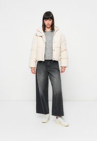 Puffy kremjakke med hette, grå strikkegenser, vide mørke jeans og hvite joggesko på en ensfarget bakgrunn.