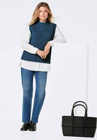 Dunkelblause gestrickte ärmellose Bluse über einem weißen Hemd, kombiniert mit hellblauen Jeans. Eine schwarze strukturierte Tragetasche mit zwei Griffen steht daneben.