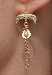 Boucles d'oreilles en forme d'oiseau en or avec un accent en goutte d'eau, présentant une pierre de couleur claire au centre, et une chaîne avec plusieurs maillons suspendue en dessous.