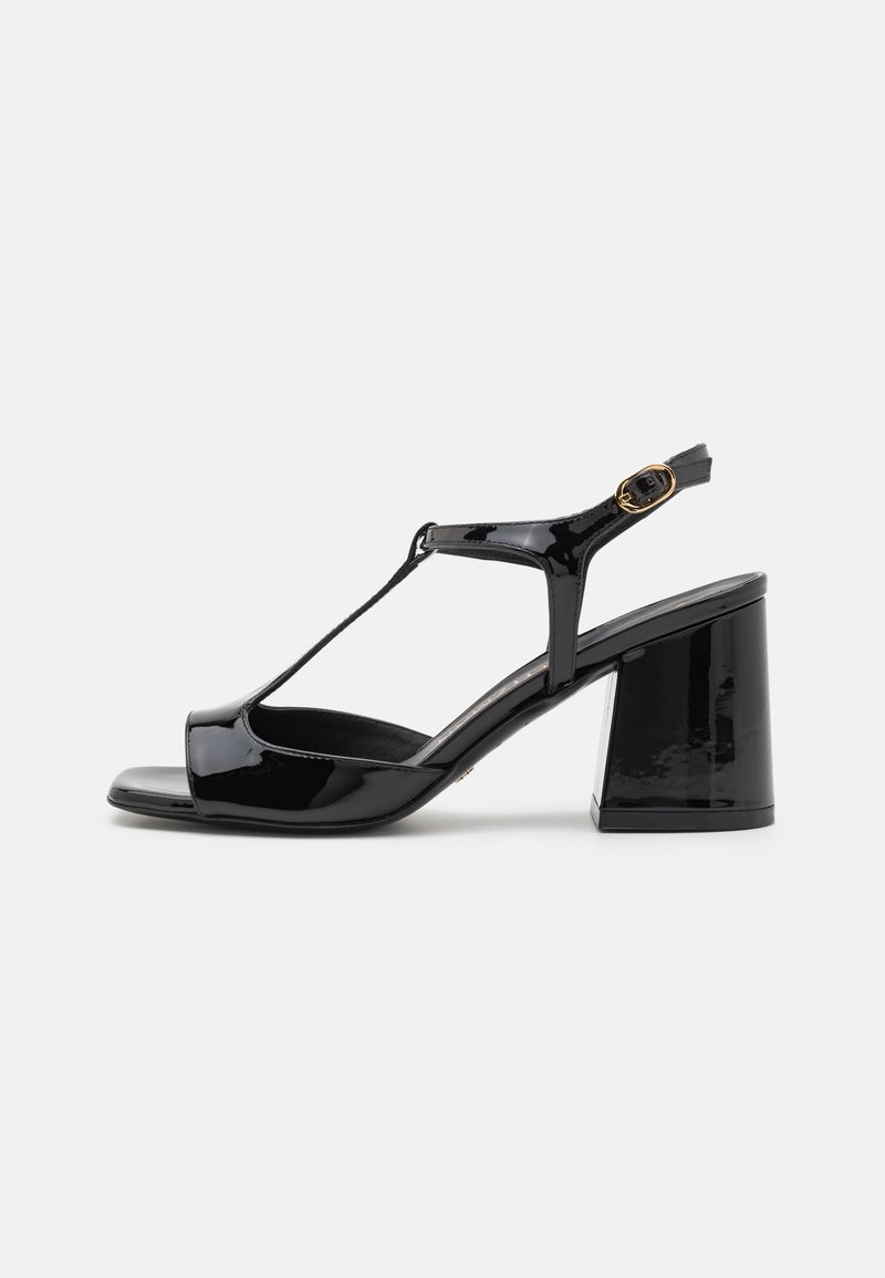 Sandale noire en cuir verni à bout ouvert avec bride arrière, design à bride en T, boucle dorée et talon bloc épais sur fond blanc.