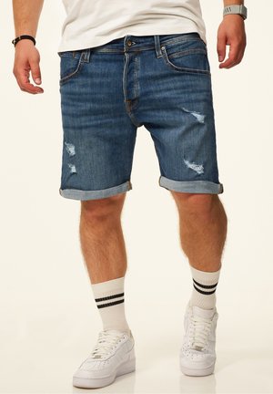 Mann trägt hochgekrempelte blaue Jeansshorts mit Rissen, weiße Sneakers, weiße Socken mit schwarzen Streifen, ein weißes T-Shirt und Armband-Accessoires.