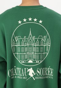 Zöld pulóver, amelyen egy nagy fehér grafika látható egy kastélyról, felette öt csillaggal, és a "CHÂTEAU SOIRÉE SKI & APRÈS SKI REFRESHMENTS" felirattal.