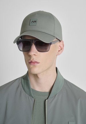 Mladić s lagano zelenom kapom s logotipom, u skladu sa svijetlo zelenom jaknom i tamnim velikim sunčanim naočalama, gleda prema naprijed s neutralnim izrazom lica.