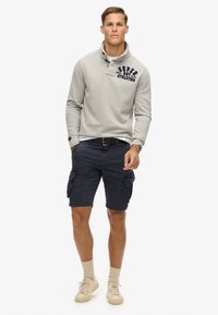 Grå fleece-pullover med krave, navy cargo-shorts, lysebrune sokker og beige sko, med et logo på brystet og lommer på shortsene.
