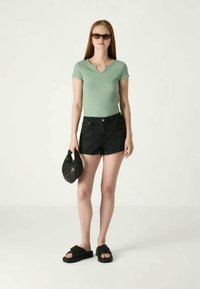 Πράσινη κοντομάνικη μπλούζα, ψηλόμεσα μαύρα τζιν σορτς, μαύρα φλατ σαμπό και μια μικρή μαύρη τσάντα, τοποθετημένα σε ελαφρύ φόντο.