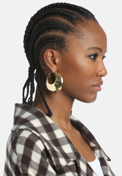 Zlatni kružni naušnici s rezbarenom dizajnom, nosi osoba s pletenom kosom, koja nosi čokoladno smeđu i bijelu flis jaknu s kariranim uzorkom.