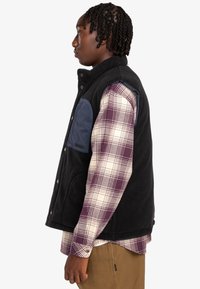 Black fleece vest with a navy nylon pocket, worn over a light plaid shirt. Features snap buttons and a relaxed fit. Textured materials.