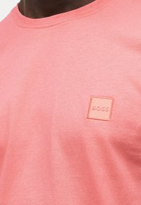Κοντινό πλάνο ενός ατόμου που φοράει ροδακινί ροζ μπλούζα με ένα μικρό τετράγωνο σημείο που φέρει την ένδειξη "BOSS" στο πάνω μέρος του στήθους.