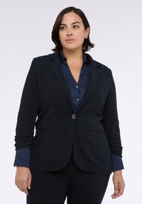 Námořnický blazer s jemně strukturovanou tkaninou, se klopami do tvaru písmene V, jednořadým zapínáním na knoflík a dvěma předními kapsami. Přes něj je přeložený odpovídající košile.