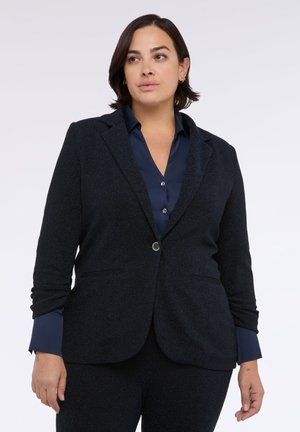 Navy blazer z subtelnie teksturowanym materiałem, klapami w kształcie litery V, zapięciem na jeden guzik i dwiema przednimi kieszeniami. Nałożony na dopasowaną koszulę.
