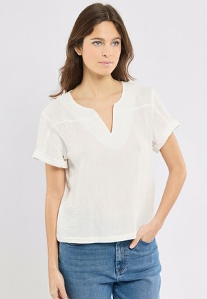 Femme aux longs cheveux bruns portant un chemisier blanc texturé à manches courtes et un jean bleu, une main dans la poche, sur un fond blanc.