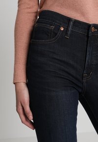 Jeans de mezclilla azul oscuro con un diseño ajustado, bolsillos frontales visibles, costuras naranjas y un botón de cobre. Arriba, un suéter de color rosa claro.